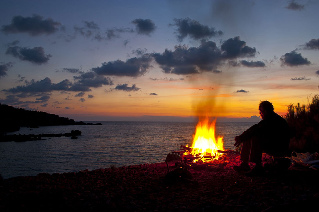 Man sits next to campfire while the sun sets on a bay.