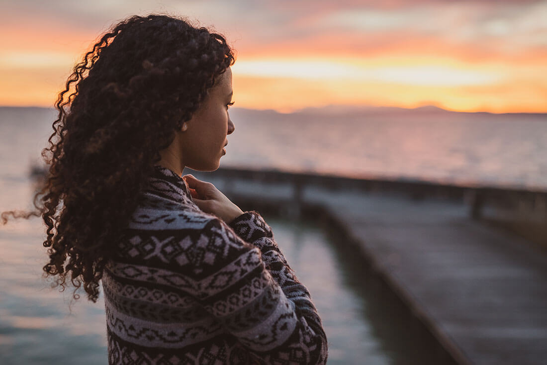 Grieving girl looks out over a lake while the sun sets.